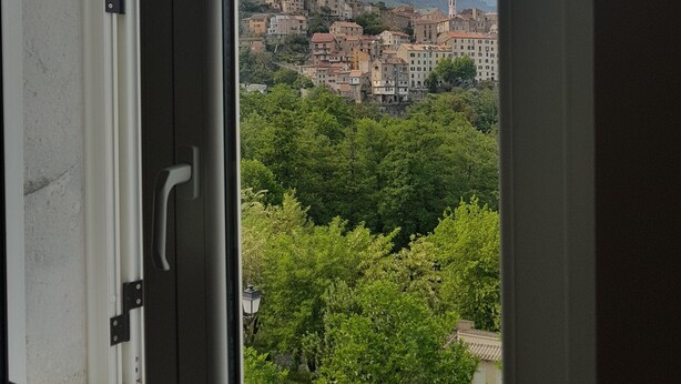 A window view of a town with buildings on a hillside, greenery, and mountains in the background.