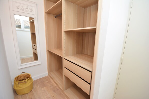 Un dressing ouvert avec des étagères et un seul tiroir, un miroir sur le côté gauche et un sac jaune posé sur le sol.