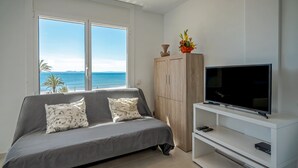 A modern bedroom with a large window, a grey sofa, a wooden cabinet, and a TV on a white stand. - Amazing apartment on the seafront in Roses Inmosantos Oasis F5 (Rosas / Roses)
