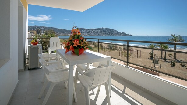 A balcony with a white table and chairs, a flower vase, and a view of the beach and mountains. - Amazing apartment on the seafront in Roses Inmosantos Oasis F5 (Rosas / Roses)