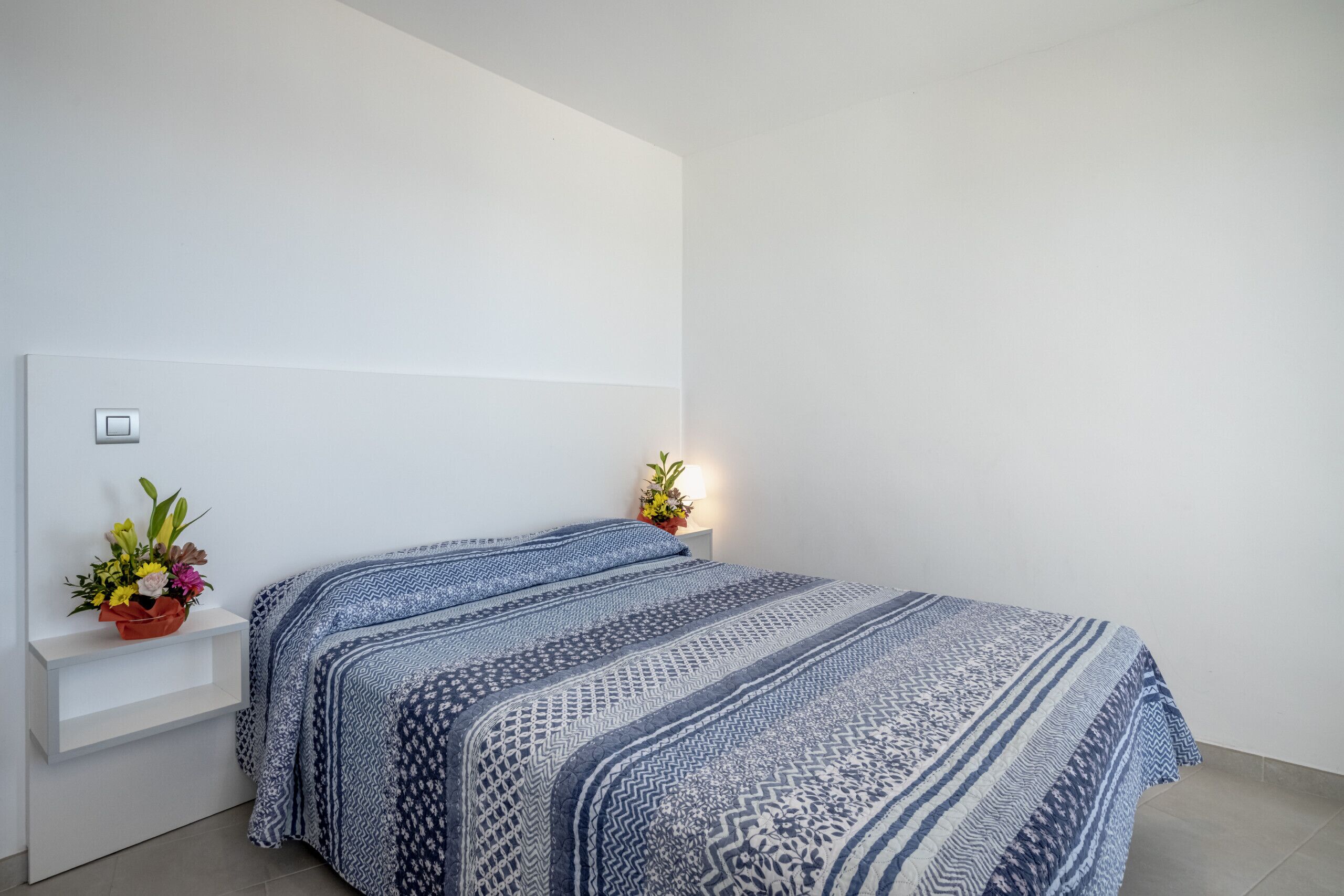 A bedroom with a bed, a nightstand, and a wall-mounted shelf with a flower arrangement.