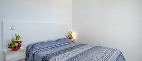 A bedroom with a bed, a nightstand, and a wall-mounted shelf with a flower arrangement.