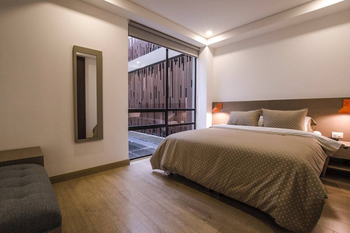 Une chambre moderne avec un grand lit, une table de chevet, un miroir et une fenêtre donnant sur une structure en bois à l’extérieur.