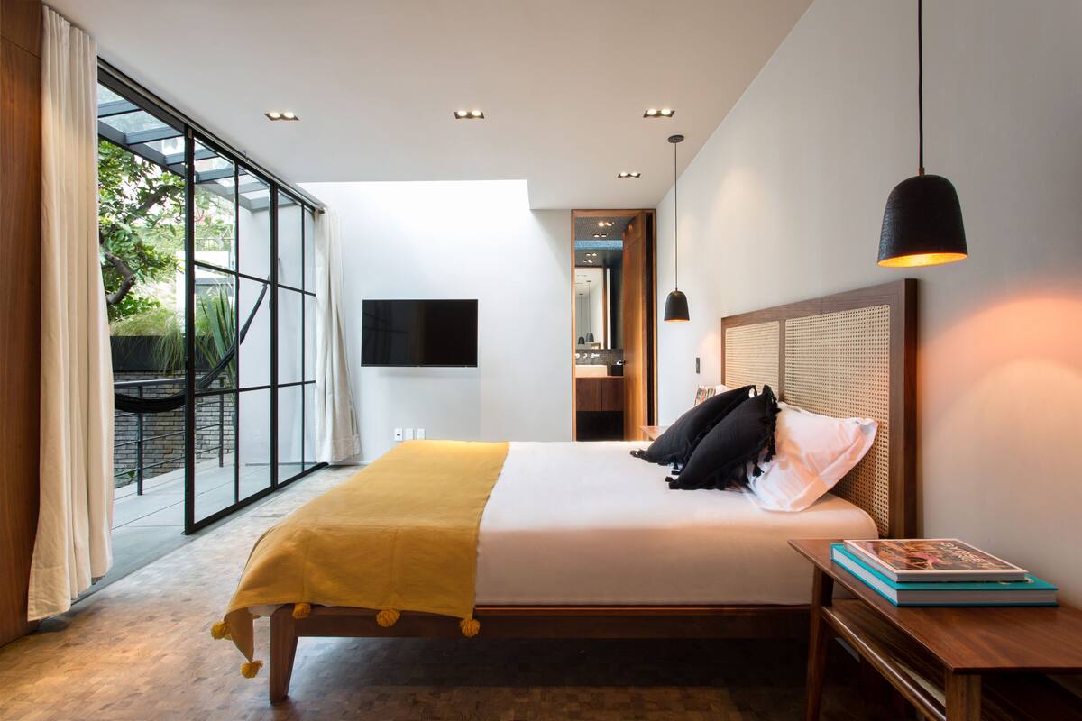 A modern bedroom with a large bed, a bedside table, a TV mounted on the wall, and a view of the outdoors through a glass door.