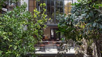 An outdoor dining area with tables and chairs, surrounded by lush greenery and a modern building with large windows.