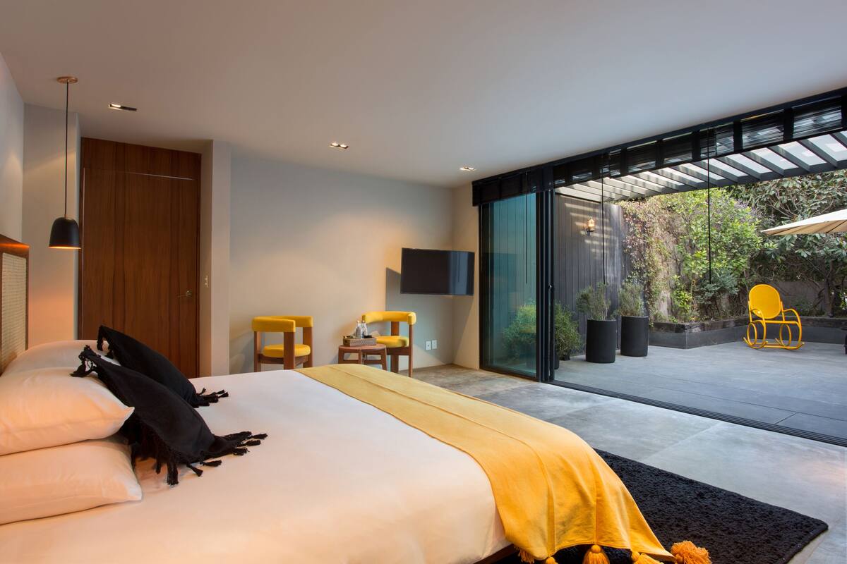 A modern hotel room with a large bed, a desk, two chairs, and a glass door leading to an outdoor area with plants and a chair.