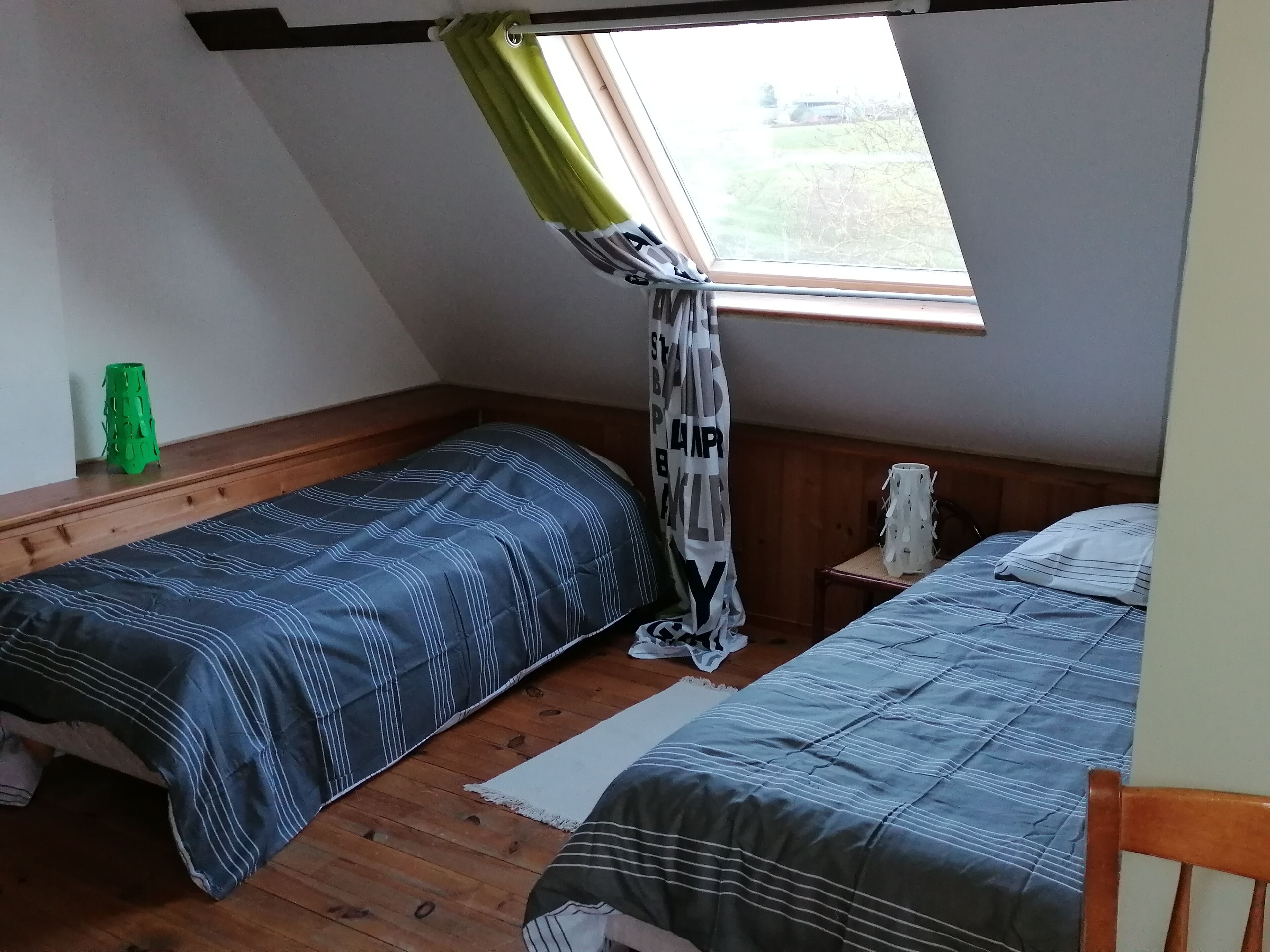 A small, cozy room with two single beds, a wooden floor, a window with a green curtain, and a small green stool.