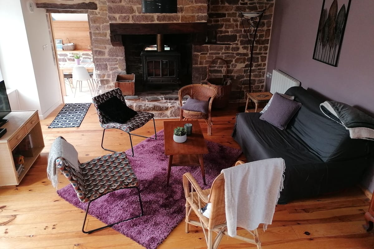 A cozy living room with a stone fireplace, wooden flooring, and a black sofa with cushions. There are two wicker chairs and a small wooden table with a plant.