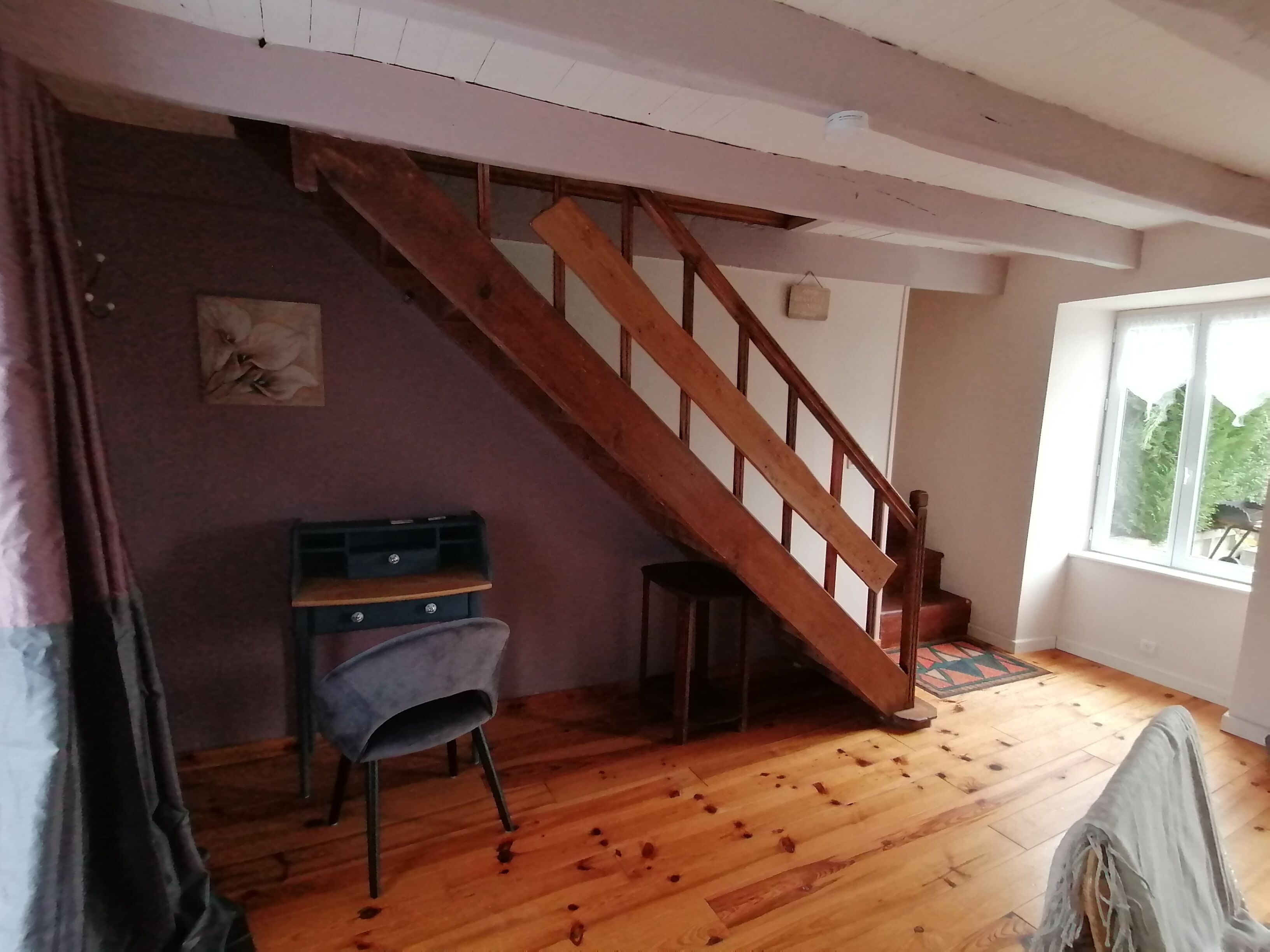 A room with a wooden staircase, a desk with drawers, a chair, and a window with a view of greenery.
