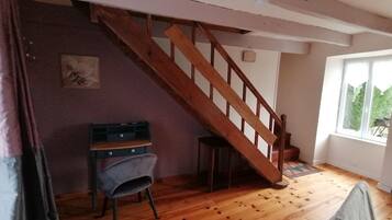 A room with a wooden staircase, a desk with drawers, a chair, and a window with a view of greenery.