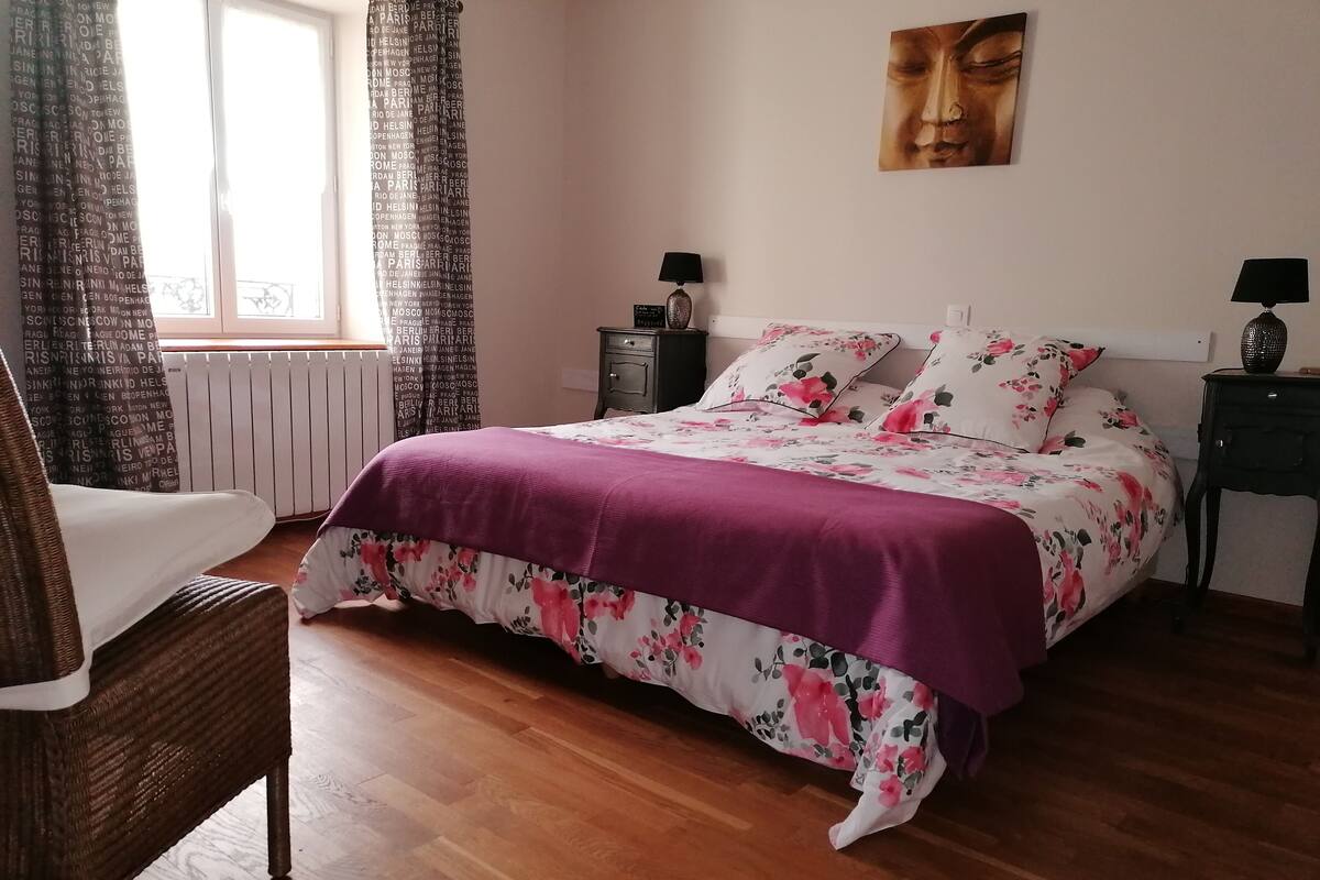 A bedroom with a bed, a chair, a nightstand, a lamp, and a window with curtains.