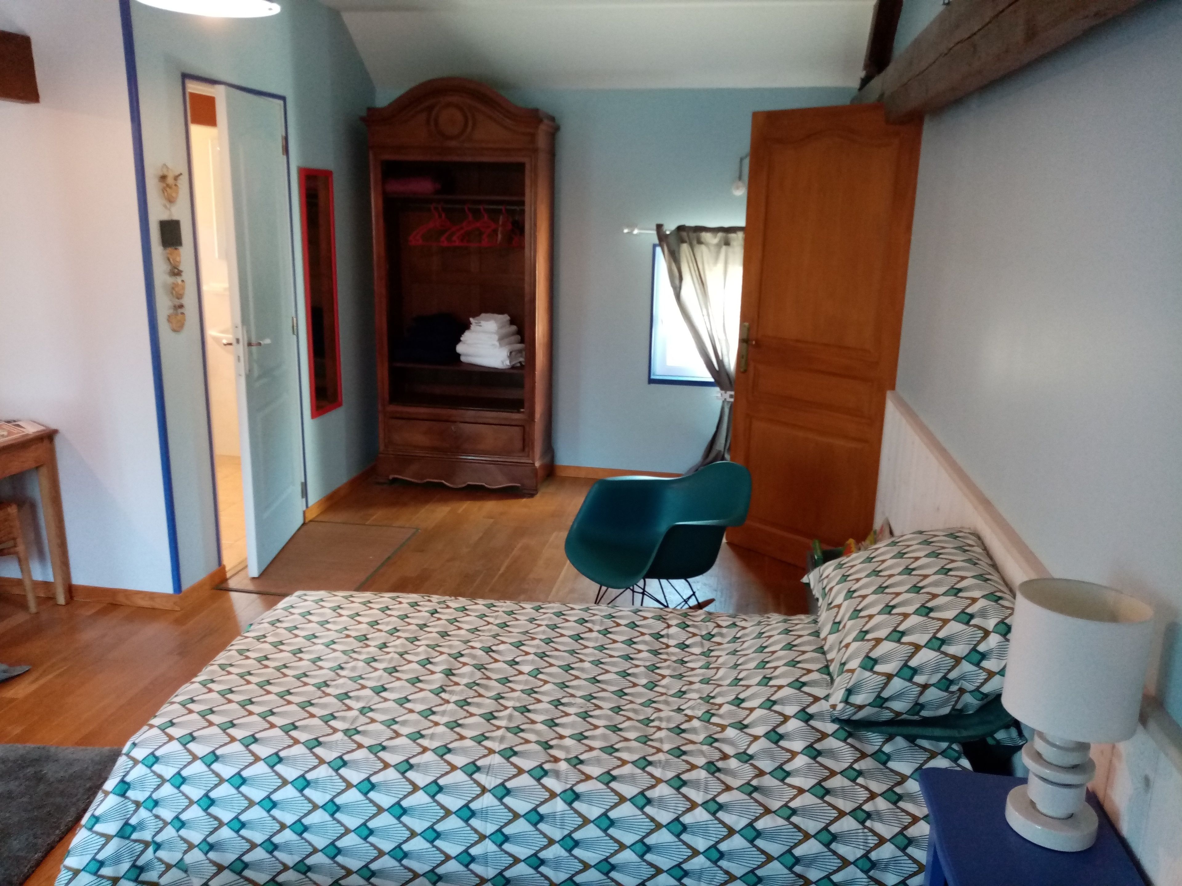Un dormitorio con una colcha estampada, un armario de madera, una silla azul y una mesita de noche con lámpara.