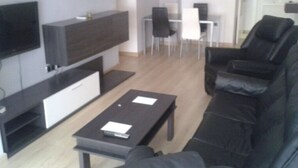 Una sala de estar moderna con un sofá negro, una mesa de centro, un televisor de pantalla plana montado en la pared y un comedor con una mesa y sillas blancas.