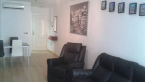 Una sala de estar con un sofá de cuero negro, un sillón de cuero negro y una mesa de centro blanca. Hay un cuadro en la pared y una puerta que da a otra habitación.