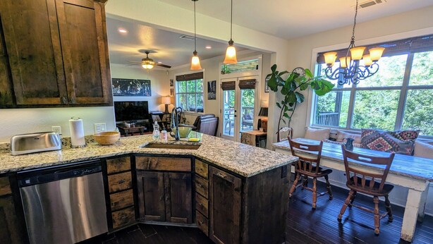 A kitchen with dark wood cabinets, granite countertops, and a dishwasher. There is a dining area with a table and chairs, and a large window providing a view of the outdoors.