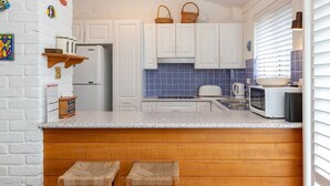 A kitchen with white cabinets, a granite countertop, a microwave, a toaster, and a sink. There are two wicker stools and a basket on the countertop. - Oceanside 20 - Sapphire (Boomerang Beach)