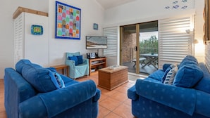 A living room with blue sofas, a patterned wall hanging, a television, and a sliding glass door leading to an outdoor patio. - Oceanside 20 - Sapphire (Boomerang Beach)
