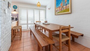 A dining area with a wooden table and benches, white brick walls, and a painting of a beach scene. - Oceanside 20 - Sapphire (Boomerang Beach)