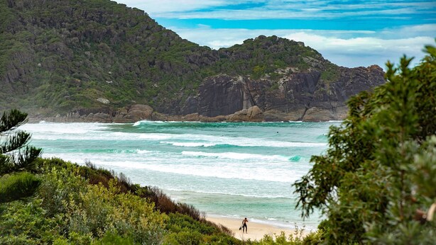 A beach with waves, a person with a surfboard, and a mountain in the background. - Oceanside 20 - Sapphire (Boomerang Beach)