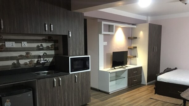 Een kleine keuken met een magnetron, koelkast en wastafel. Er staat een bed met een houten hoofdeinde en een klein tafeltje.