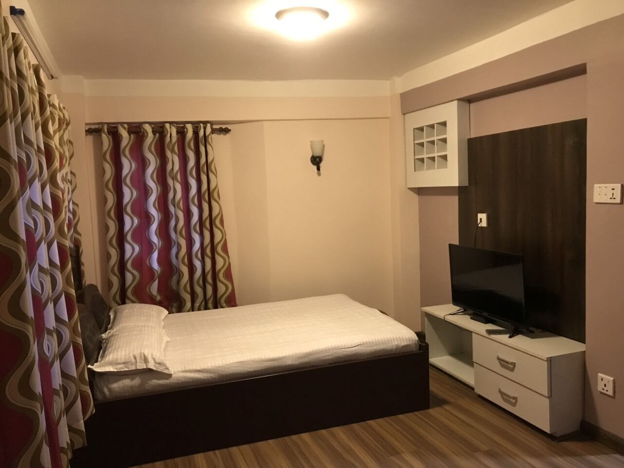 Un dormitorio con una cama, un televisor sobre un mueble, una lámpara de pared y una cortina.