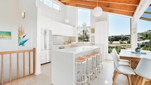 Een moderne keuken met witte kasten, een centraal eiland met krukken, grote ramen en uitzicht op de omgeving.