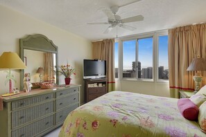Habitación de hotel con cama, televisor, cómoda, ventilador de techo y ventana con vista a la ciudad.