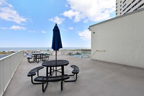 Terraço no telhado com mesas de piquenique e um guarda-sol azul, com vista para uma praia e um estacionamento.
