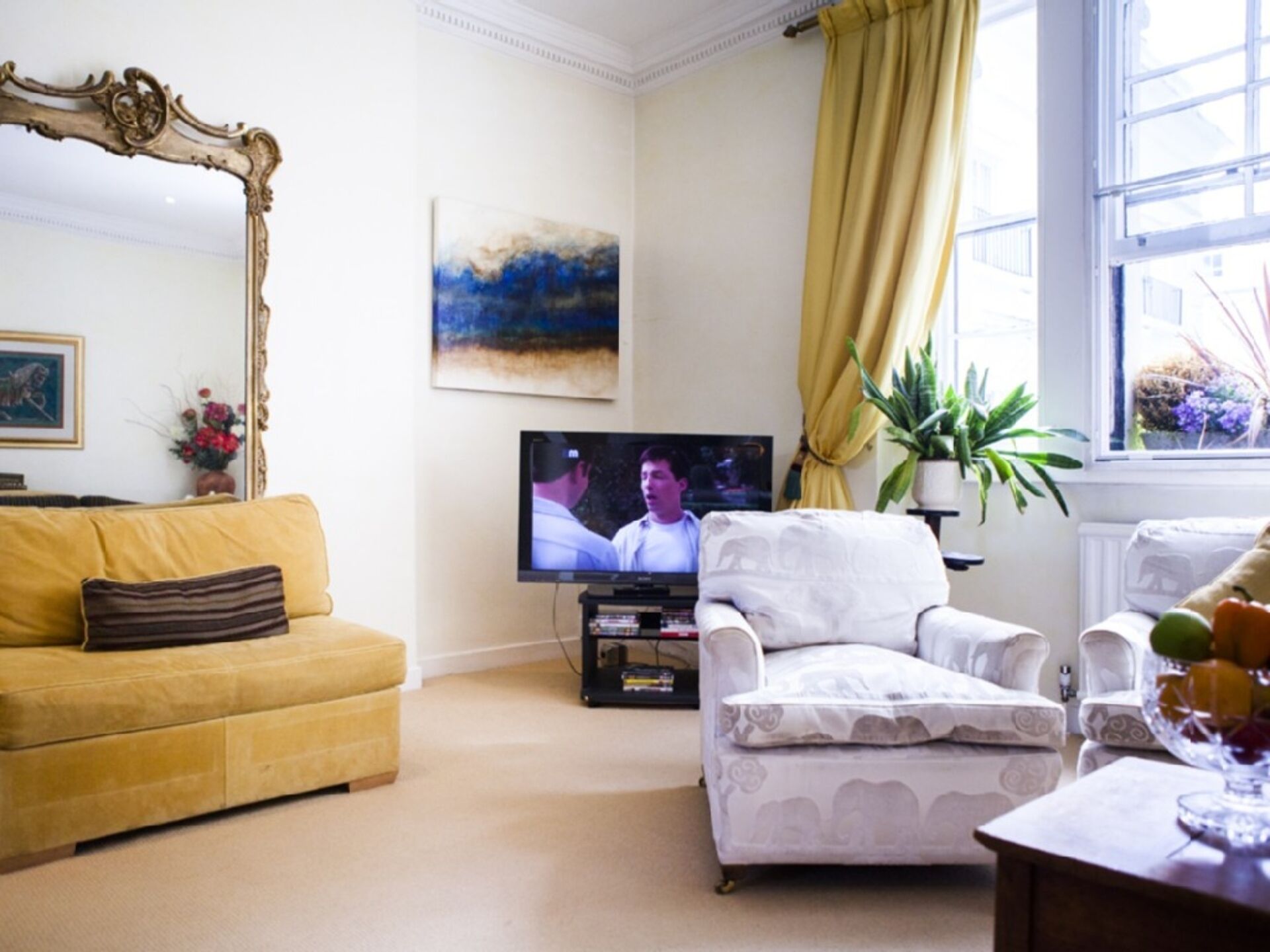 A living room with a yellow sofa, a white armchair, a television, and a painting on the wall.