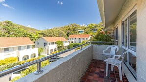 Un balcon doté d’une rambarde en métal, de deux chaises en plastique blanches et offrant une vue sur des maisons aux toits rouges, avec une végétation luxuriante en arrière-plan.