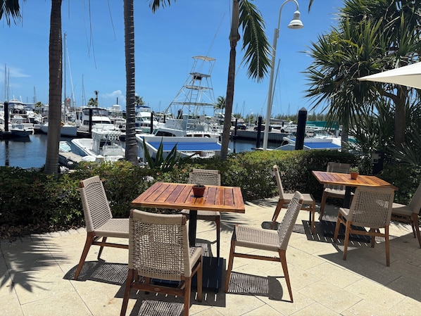 A patio with wooden tables and wicker chairs, overlooking a marina with boats and a clear blue sky.