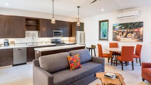 A modern kitchen with dark wooden cabinets, a microwave, and a refrigerator. There is a dining area with orange chairs and a table. A gray sofa with colorful pillows is in the living room, and a wooden coffee table is in the center. - Casa Del Gallo 2 BR by Casago (Loreto)