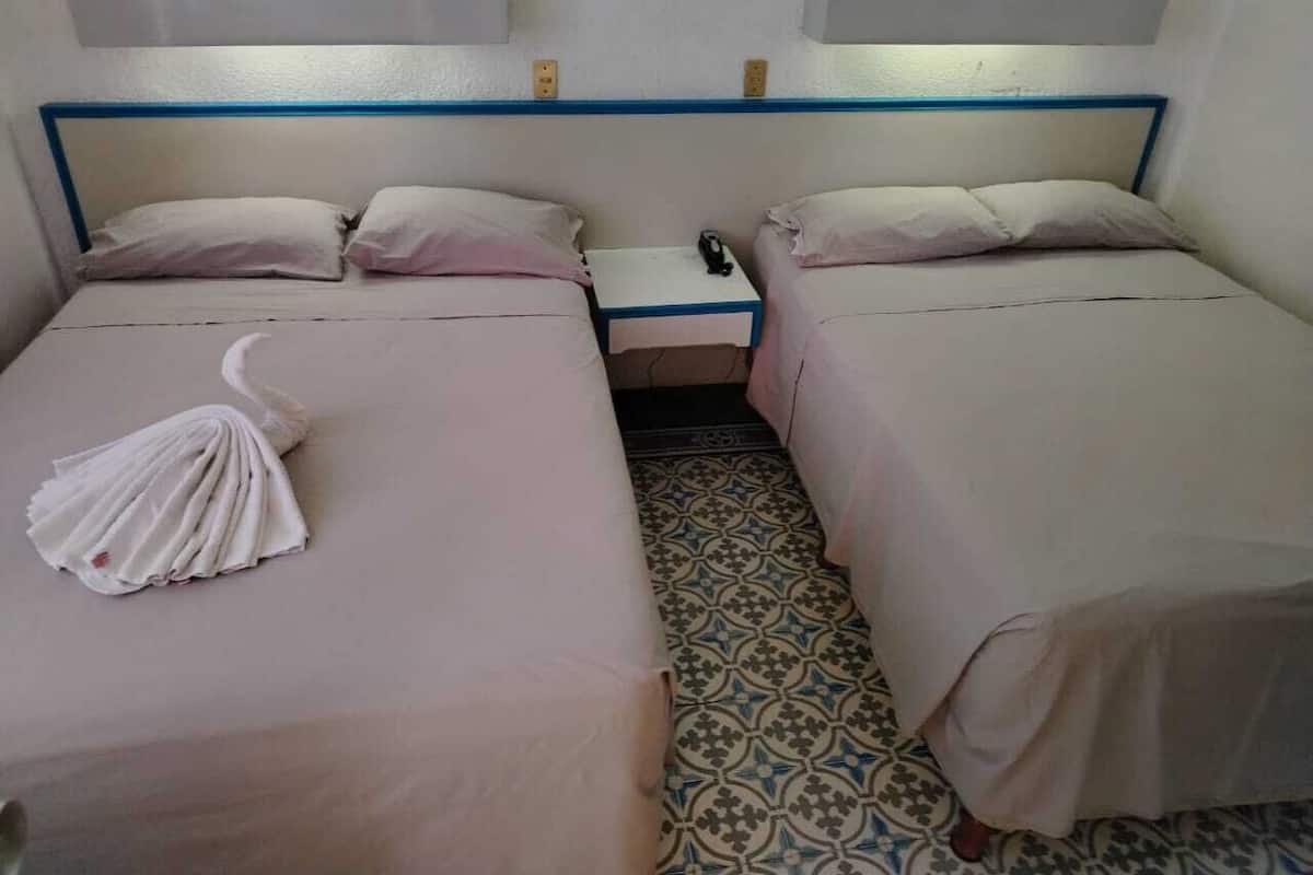 A hotel room with two beds, each with a pillow and a folded towel. There is a nightstand between the beds with a phone and a lamp. The room has a patterned floor and a white wall with a blue trim.