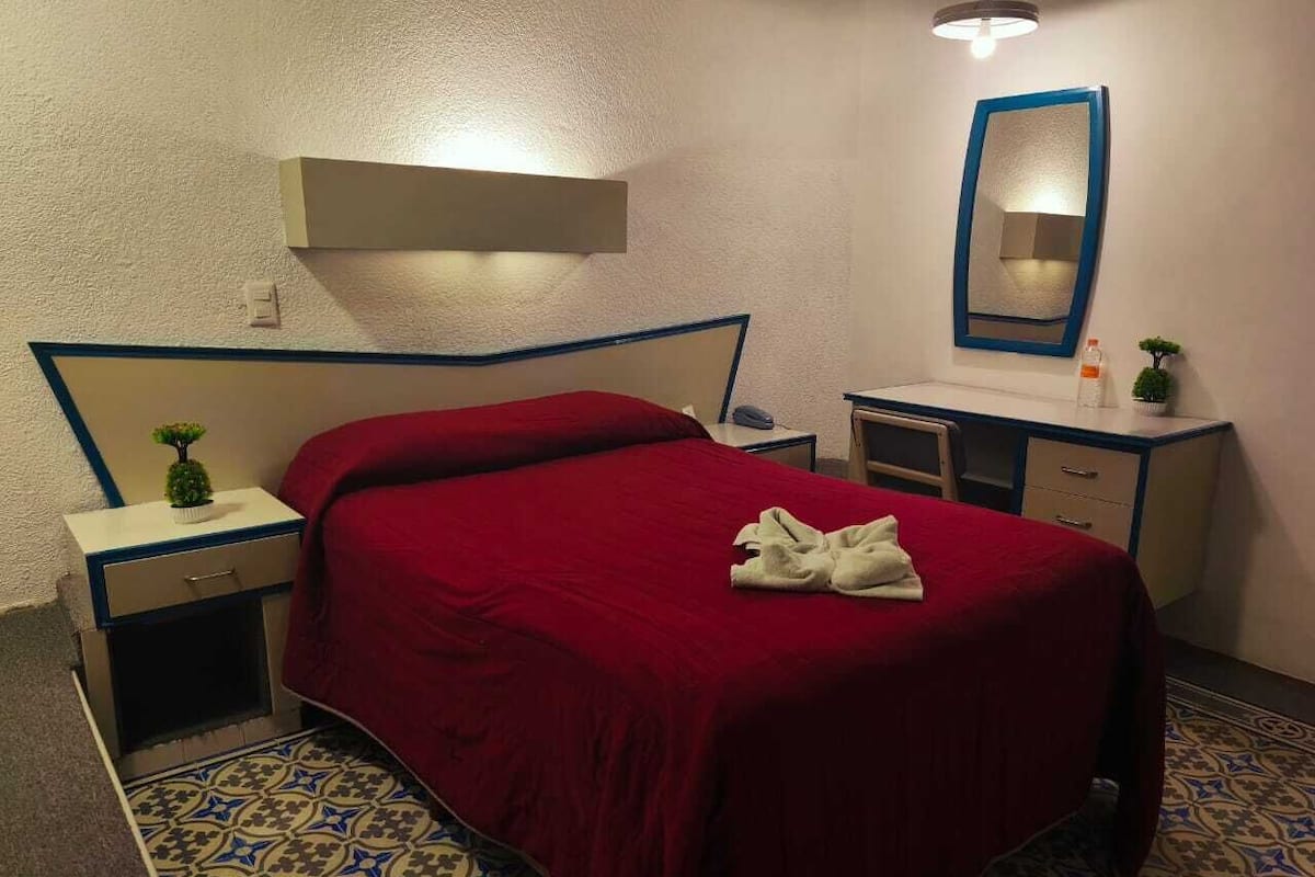 A hotel room with a bed, a nightstand, a dresser, a mirror, and a small plant.