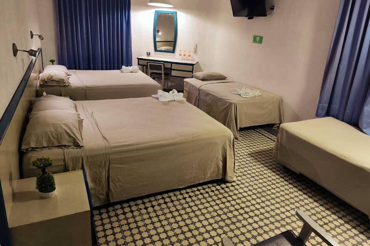 A hotel room with three beds, a desk with a mirror, a TV mounted on the wall, and a patterned floor.