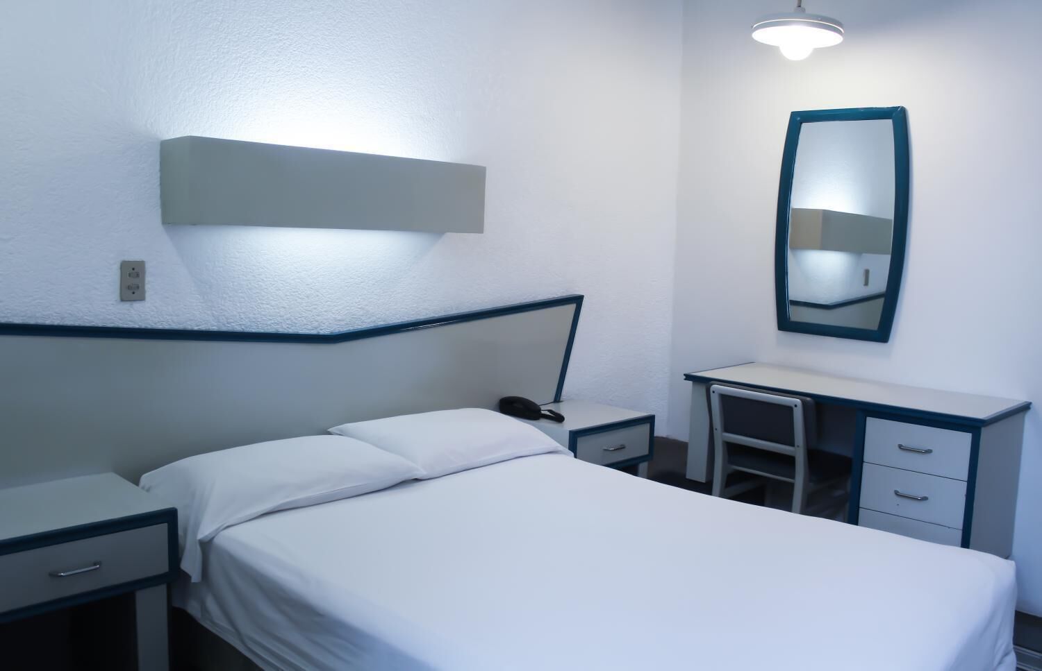 A hotel room with a single bed, a bedside table, a desk with drawers, a mirror, and a light fixture above the desk.