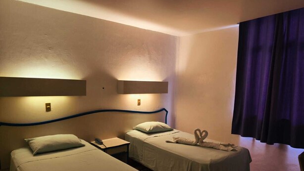 Quarto de hotel com duas camas de solteiro, uma mesinha de cabeceira com abajur, telefone e uma toalha decorativa em formato de cisne sobre uma das camas.