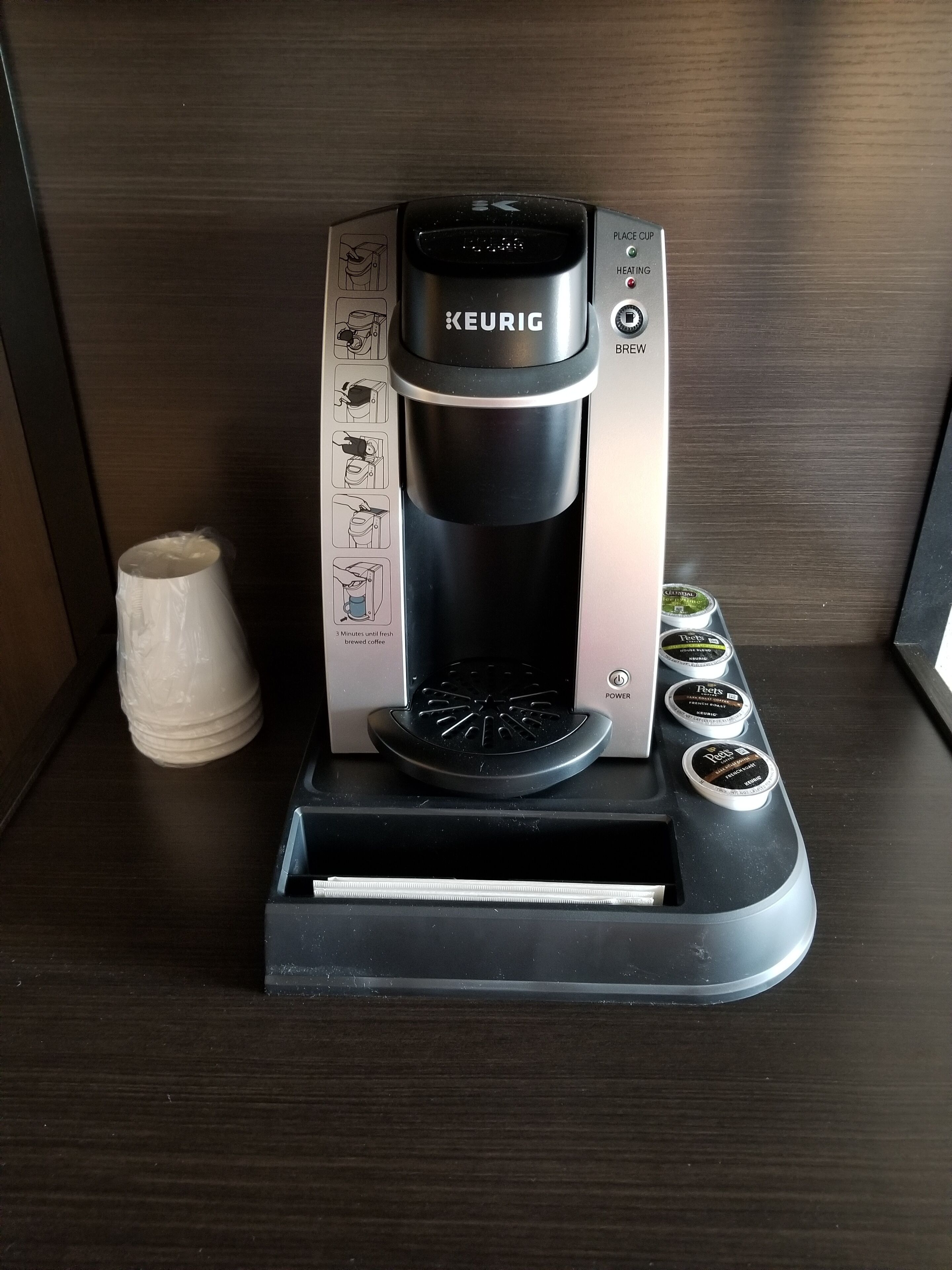 A Keurig coffee maker with a drip tray, a coffee pod holder, and a stack of paper cups on a wooden surface.