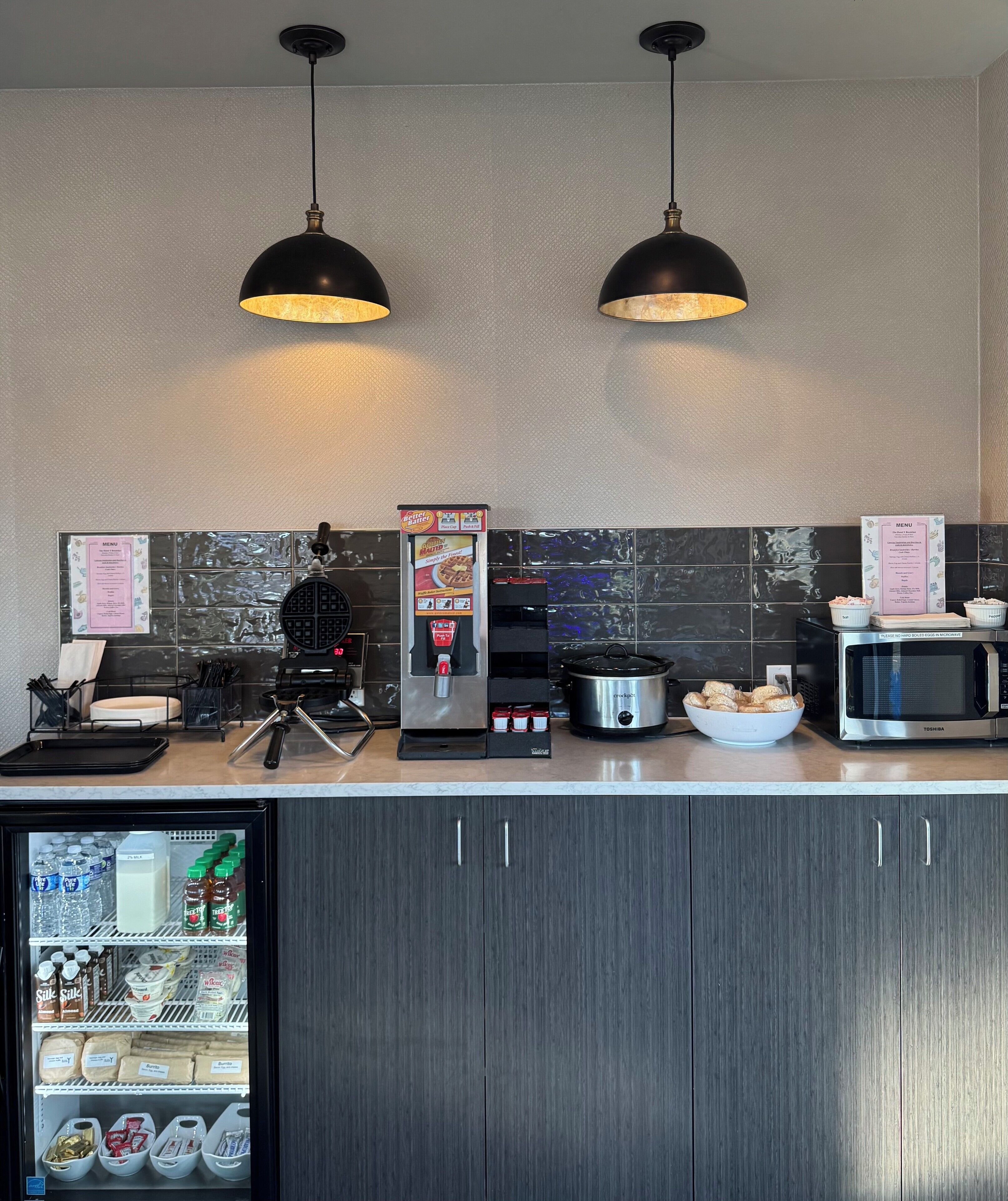 A modern kitchen with a refrigerator, microwave, coffee machine, and a food display.
