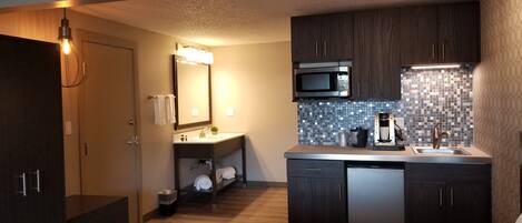A compact hotel room with a bed, a kitchenette, a bathroom, and a small seating area.
