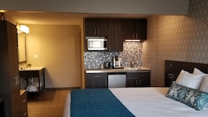A hotel room with a large bed, a kitchenette with a microwave, and a bathroom with a sink and mirror. - The Hotel Y, a Dazzler Select by Wyndham (Yakima)