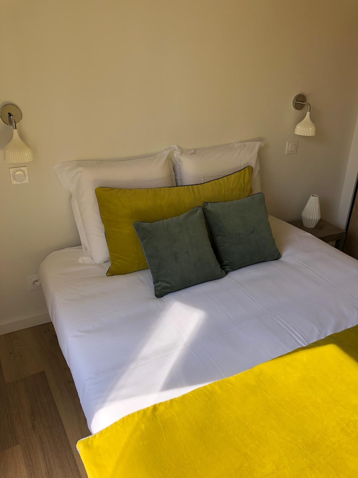 Un lit avec une literie blanche et deux oreillers, un jaune et un vert, dans une pièce dotée d’un plancher en bois et d’un mur blanc.
