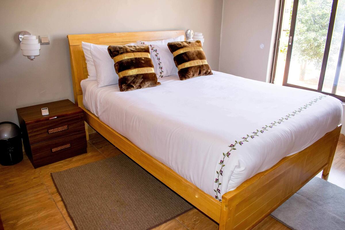 A wooden bed with white bedding and two decorative pillows, a bedside table with a lamp, and a view of greenery through a window.