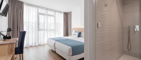 Habitación de hotel con cama, escritorio, silla, televisor y zona de ducha.
