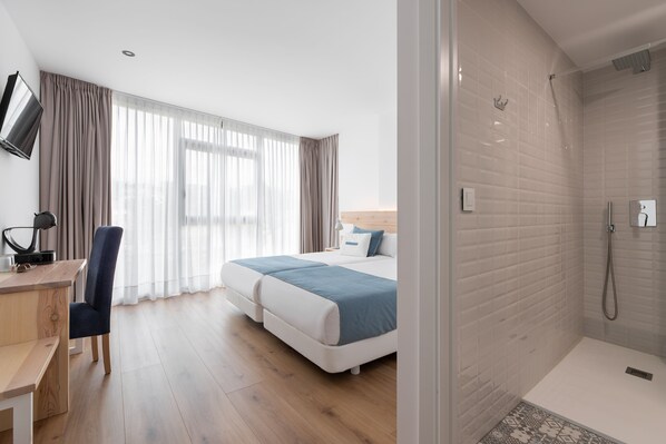 A hotel room with a bed, a desk, a chair, a TV, and a shower area.