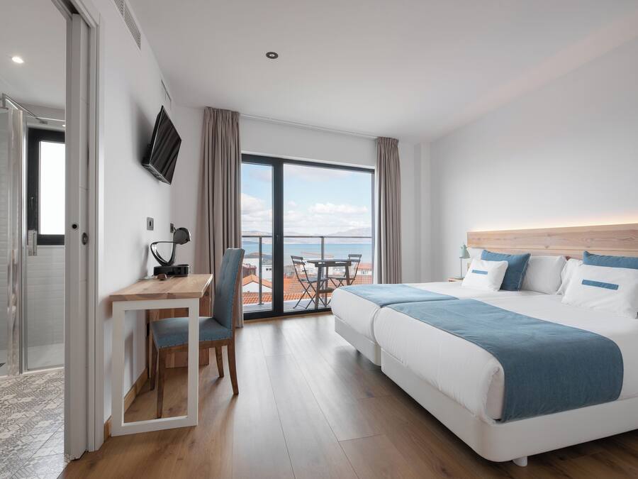 Habitación de hotel con cama, escritorio, silla y un gran ventanal con vista al mar.