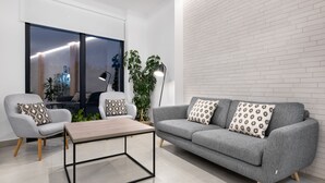 A modern living room with a grey sofa, a grey armchair, and a wooden coffee table. There is a large window with a view of the city, a potted plant, and a floor lamp. - Hotel Costa da Morte (Fisterra)