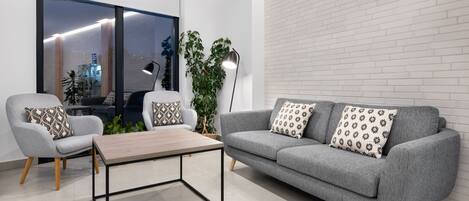 A modern living room with a grey sofa, a grey armchair, and a wooden coffee table. There is a large window with a view of the city, a potted plant, and a floor lamp.