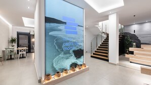A modern hotel lobby with a large digital screen displaying 'Hotel Costa da Morte', a staircase, and a seating area with wooden benches. - Hotel Costa da Morte (Fisterra)