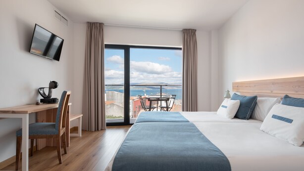 A hotel room with a bed, a desk, a chair, a TV, and a balcony with a view of the sea. - Hotel Costa da Morte (Fisterra)